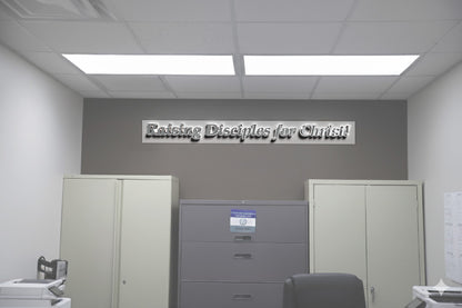 Office interior with metallic 'Raising Disciples for Christ!' wall sign above gray filing cabinet and beige cabinets.
