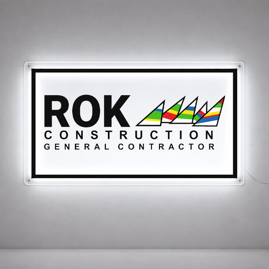 Illuminated sign on gray wall: bold ROK, CONSTRUCTION GENERAL CONTRACTOR text and colorful striped triangular sail logo