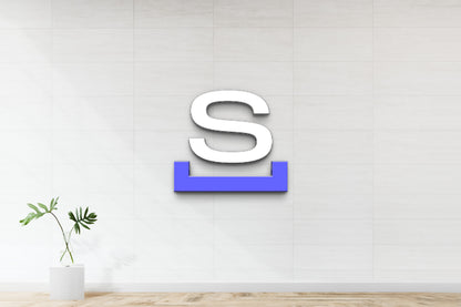 White 3D letter S with blue underline mounted on white tiled wall above glass vase holding monstera leaves.