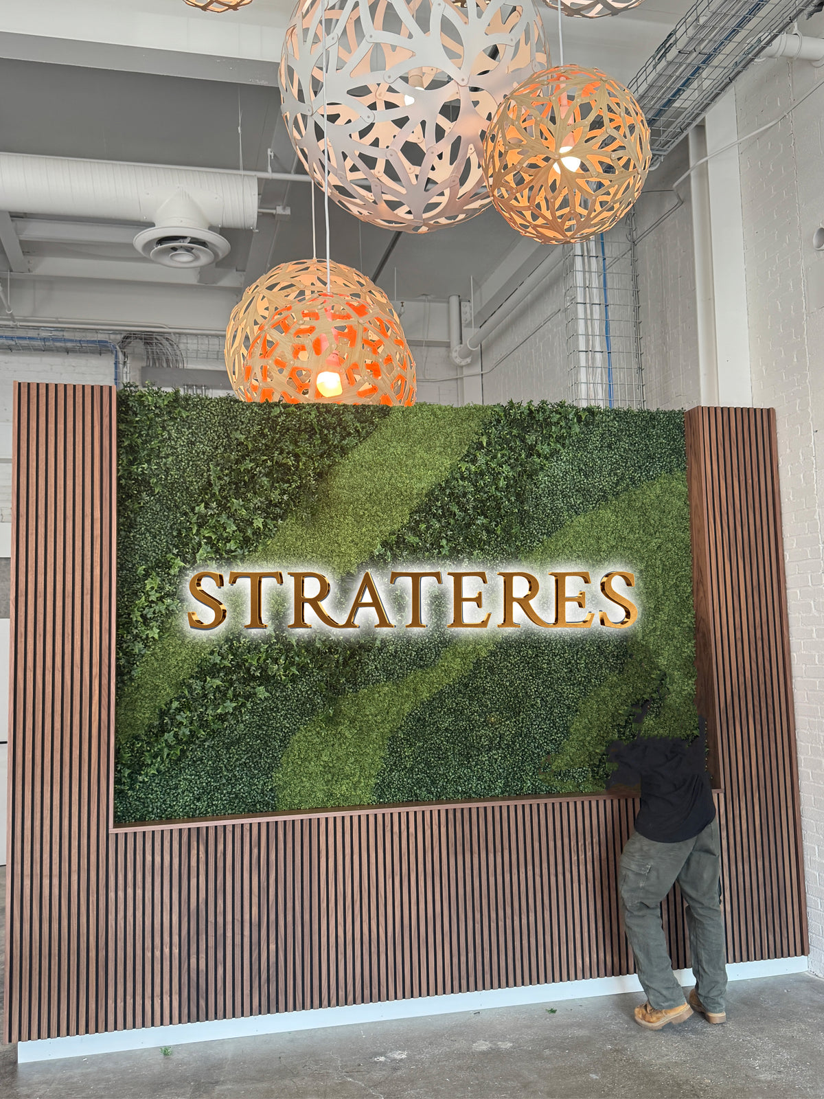 Moss vertical garden sign reading 'STRATERES' with gold letters, wood slat frame, glowing wooden orb pendant lights.