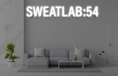 Glowing neon 'SWEATLAB:54' above a gray living room with sectional sofa, glass coffee table, potted bonsai, marble wall.