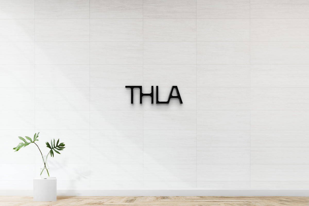 White tiled wall with black THLA logo centered, glass vase of green tropical leaves on white pedestal, light wood floor.