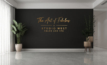 The Art of Fabulous Studio West 3D gold metal script logo on black accent wall, nonlit dimensional letters in luxe salon spa interior.