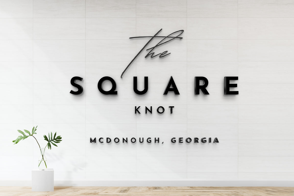 Black 3D signage reading 'The SQUARE KNOT' mounted on white tile wall with a glass vase and green plant left.