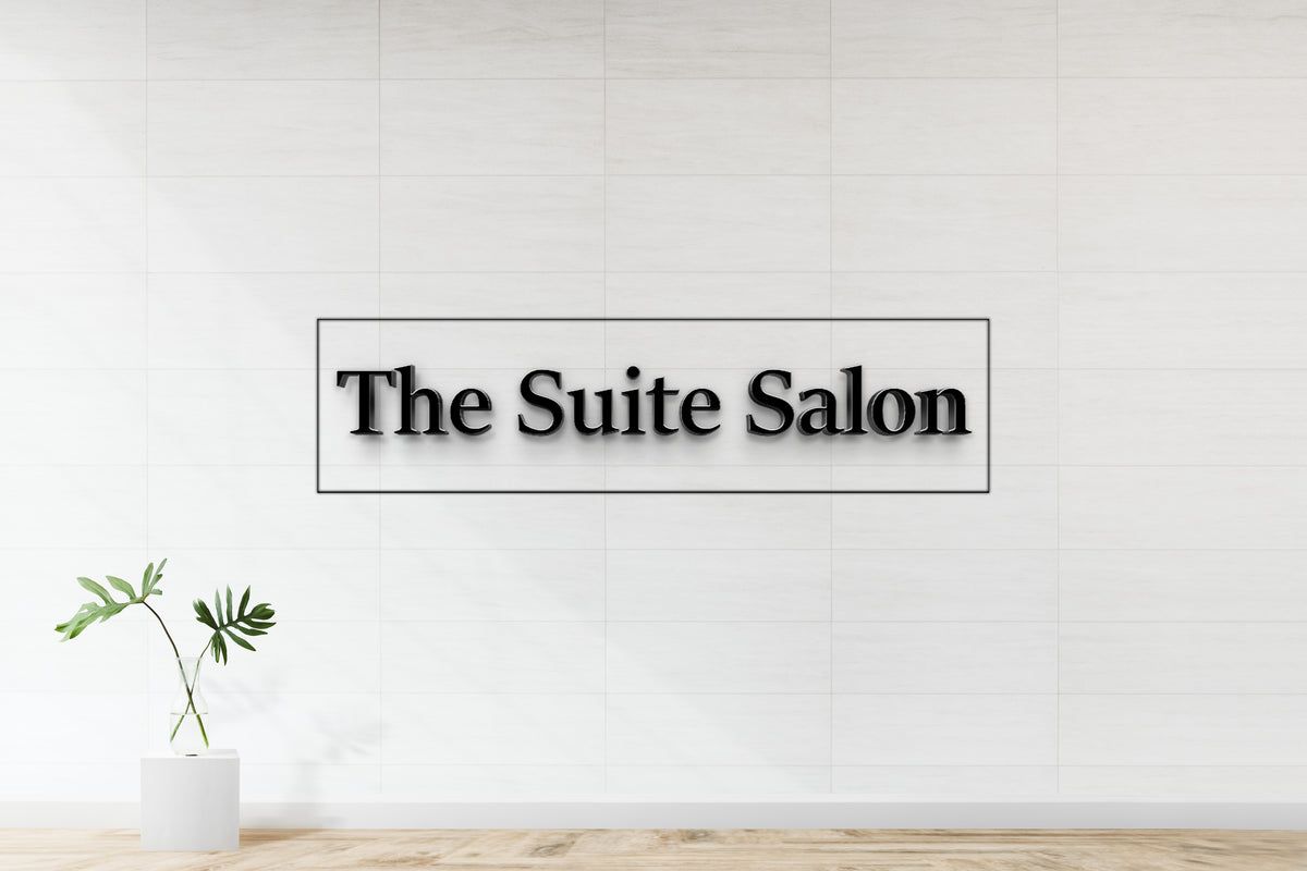 Black 3D 'The Suite Salon' sign in thin rectangle on white tiled wall above glass vase with monstera leaves.