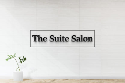 Black 3D 'The Suite Salon' sign in thin rectangle on white tiled wall above glass vase with monstera leaves.