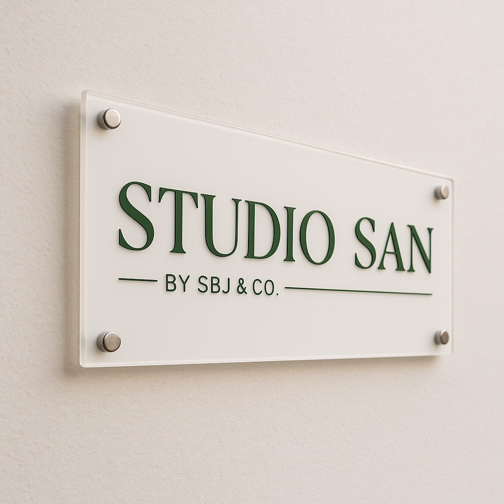 Wall-mounted acrylic sign with green 'STUDIO SAN' lettering and 'BY SBJ & CO.' text on beige wall with metal standoffs.
