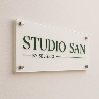 Wall-mounted acrylic sign with green 'STUDIO SAN' lettering and 'BY SBJ & CO.' text on beige wall with metal standoffs.