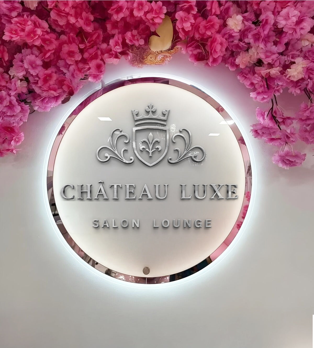 Illuminated round white sign reading 'CHÂTEAU LUXE SALON LOUNGE' with silver crest and pink blossom decor.