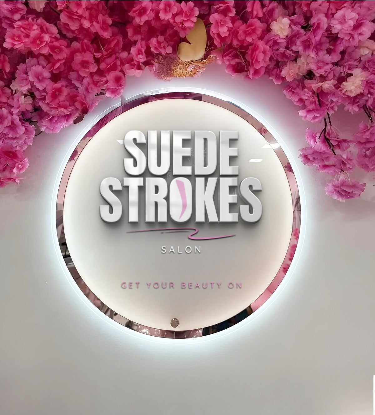 Round illuminated salon mirror sign reading 'SUEDE STROKES Salon' surrounded by pink flowers and white wall.