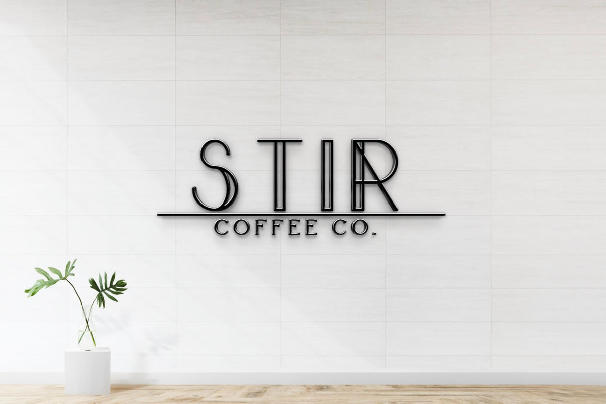 White tiled wall with black metal STIR sign, 'COFFEE CO.' underneath; glass vase with green leaves on white pedestal.