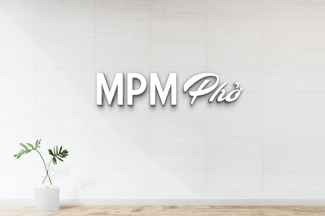 White tiled wall with raised white 3D sign reading "MPM Phở" and small potted monstera leaf on wooden floor.