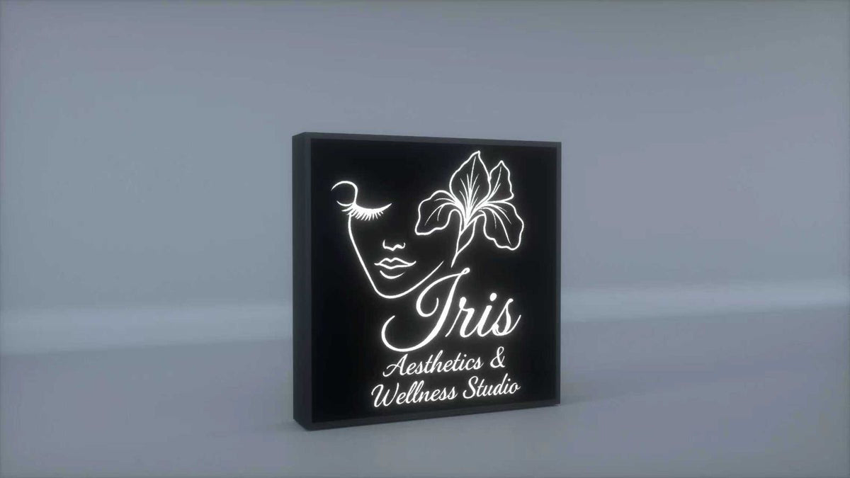 Black illuminated square sign with white line-art woman, iris flower and 'Iris Aesthetics & Wellness Studio' on gray.
