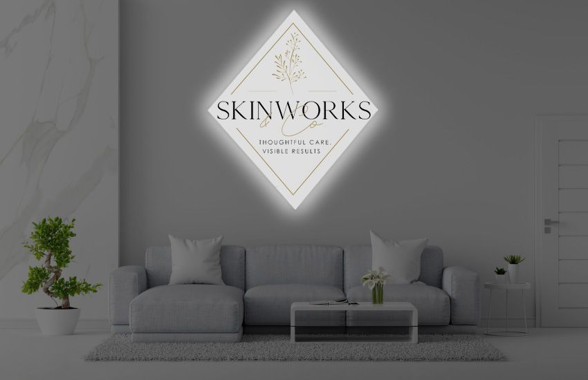 Illuminated diamond SKINWORKS logo sign with gold detailing above gray sofa in minimalist lounge with potted plants.
