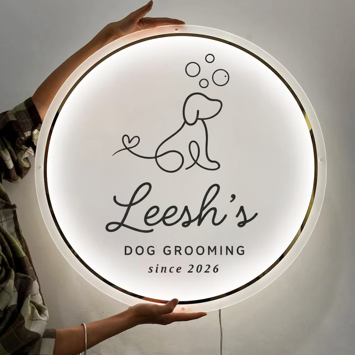 Round illuminated white sign held by hands, black line-drawn dog and bubbles, 'Leesh's DOG GROOMING since 2026'.