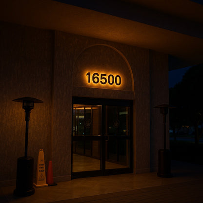 An illuminated building entrance at night with glowing address 16500 above glass doors, patio heaters and caution cone.