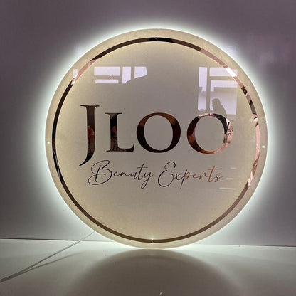 Illuminated round JLOO Beauty Experts salon sign in rose gold metallic lettering on frosted glass with halo light.