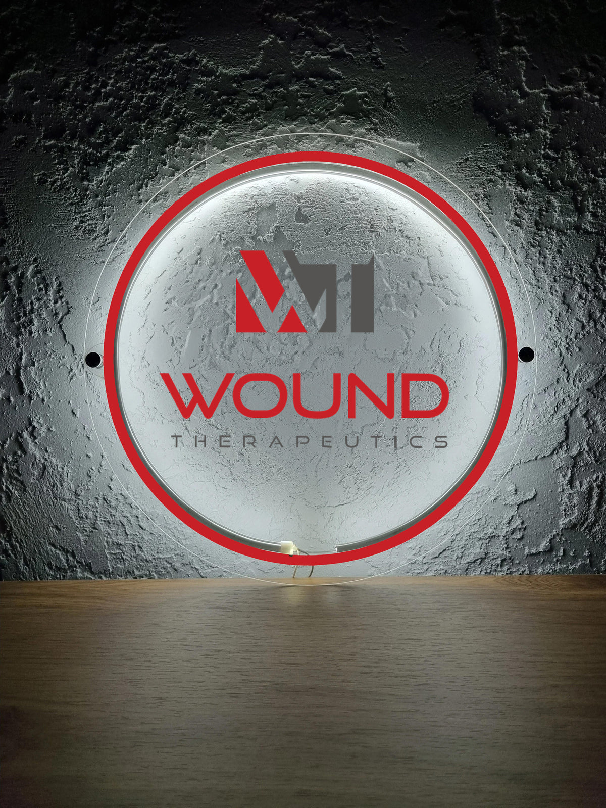 Round illuminated sign with red outer ring, grey M logo and red WOUND THERAPEUTICS lettering on dark textured wall.