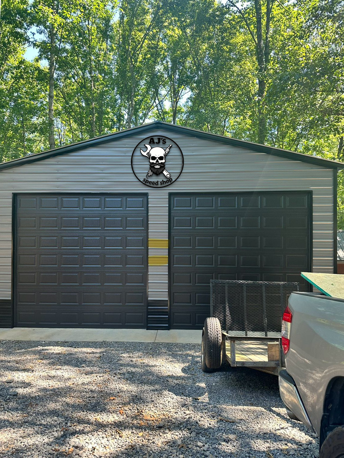 Two-bay metal garage with dark gray doors and AJ's skull logo above, pickup and trailer on gravel driveway.