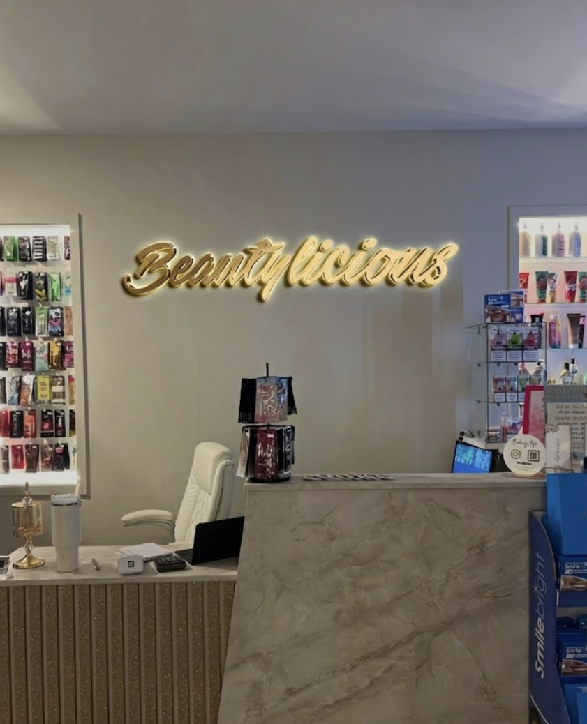 Beauty reception area with gold illuminated cursive 'Beautivicious' wall sign, marble desk, product display shelves.