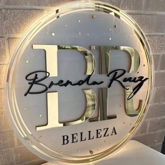 Illuminated round gold-acrylic salon sign with BR monogram, black 'Brenda Ruiz' script and 'BELLEZA' mounted on brick.