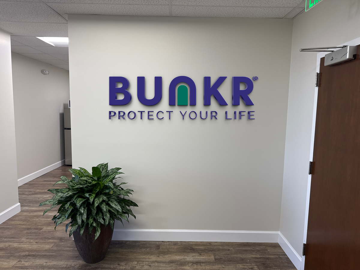 Office interior wall sign reading 'BUNKR PROTECT YOUR LIFE' purple letters with green arched U, potted plant.
