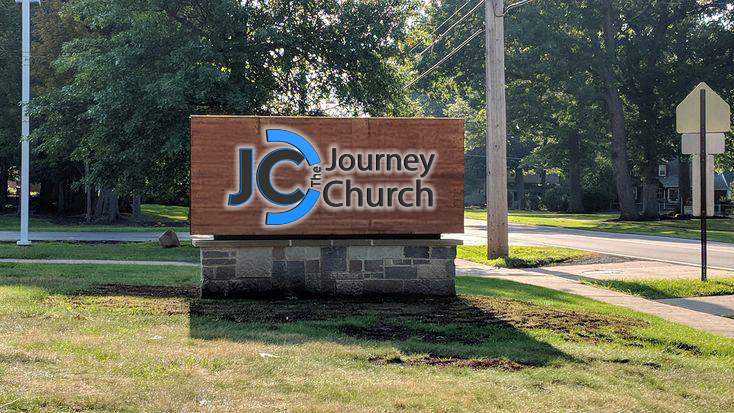 A wooden roadside sign reading 'The Journey Church' with blue JC logo mounted on a stone base by a street.