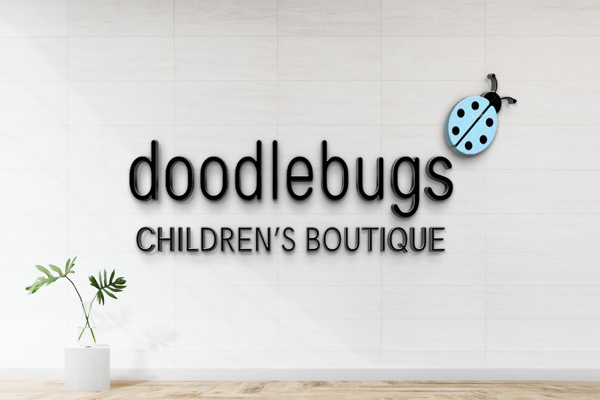 Doodlebugs children's boutique sign: black 3D letters on white tiled wall, blue ladybug logo and glass vase with leaves.