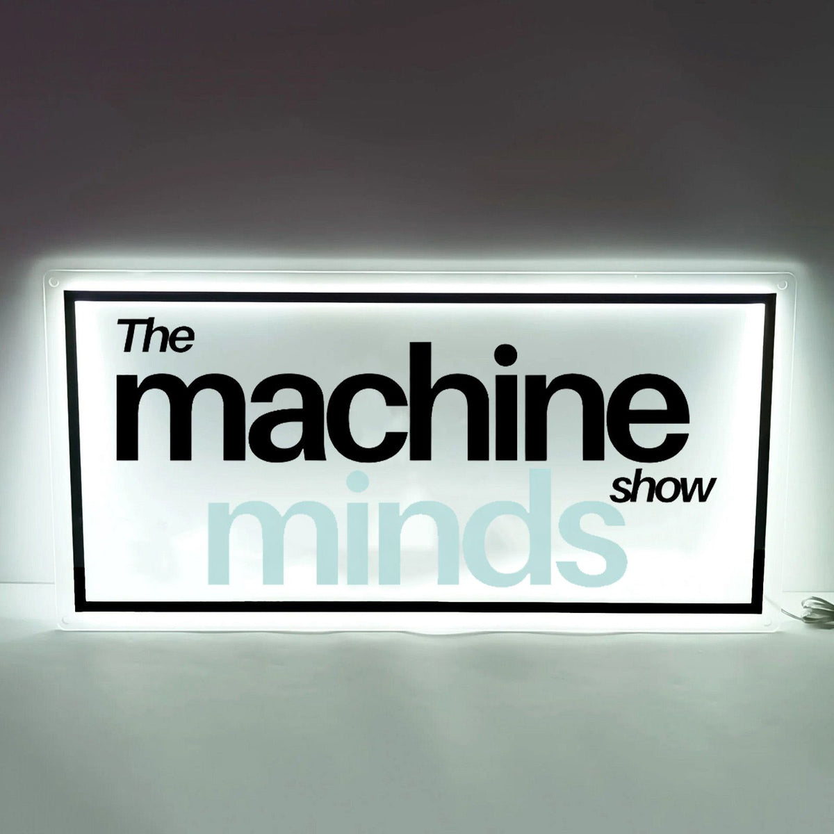Illuminated white lightbox sign with black border displaying 'The machine show' and pale blue 'minds' text.