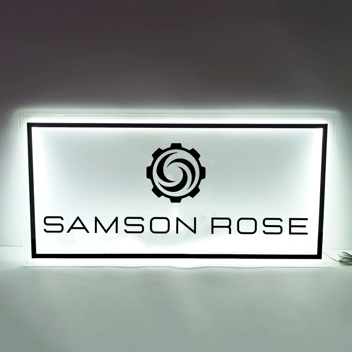Backlit white rectangular sign with black border, gear swirl logo above large black text reading 'SAMSON ROSE'.