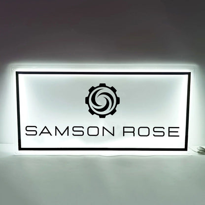 Backlit white rectangular sign with black border, gear swirl logo above large black text reading 'SAMSON ROSE'.