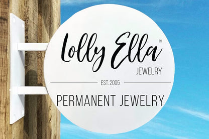 White round Lolly Ella Jewelry sign reading PERMANENT JEWELRY and EST. 2005 mounted on wooden post against blue sky.