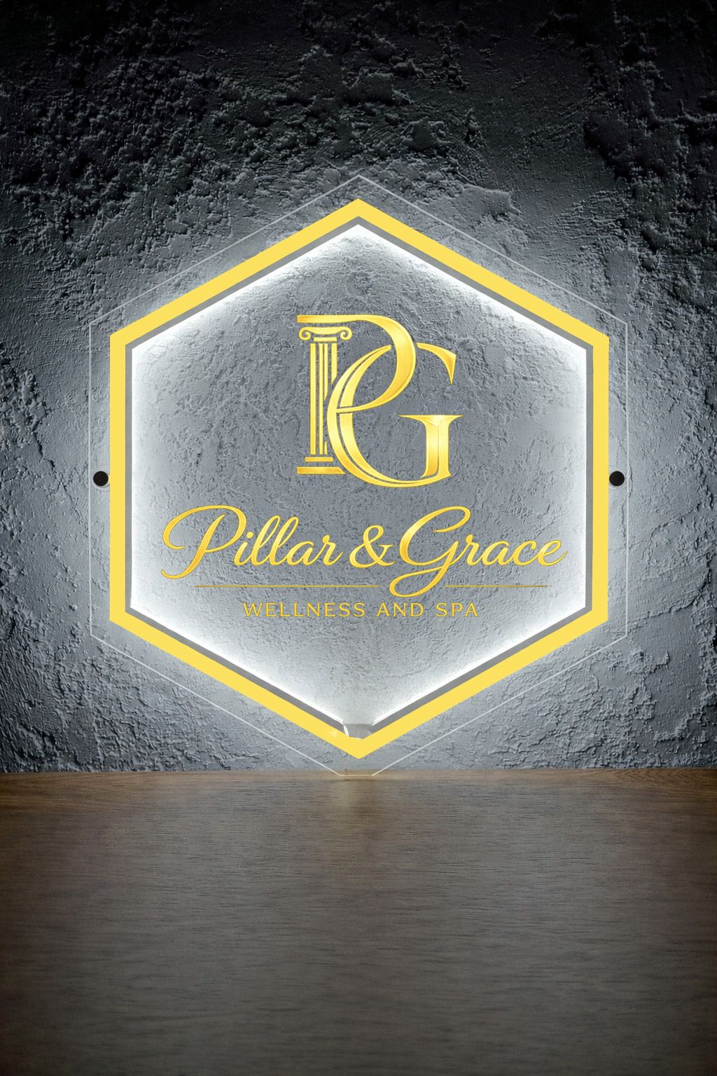 Illuminated hexagonal acrylic sign with gold PG monogram and Pillar & Grace Wellness and Spa on textured gray wall.