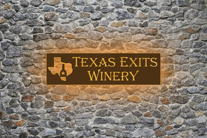 Brown rectangular sign with gold text and Texas map wine-bottle logo saying 'TEXAS EXITS WINERY' on gray stone wall.