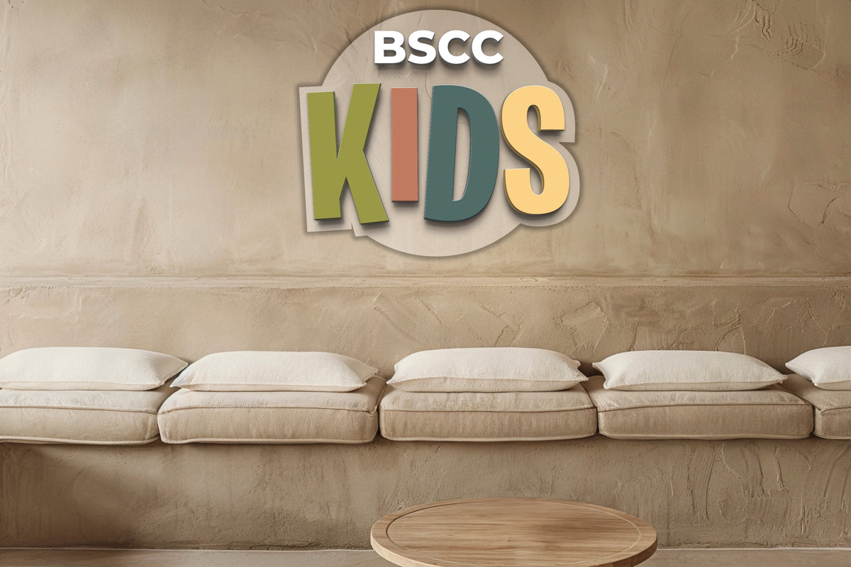 Beige plaster wall with 'BSCC KIDS' wall logo in olive, rust, teal and yellow above beige bench and round wooden table.