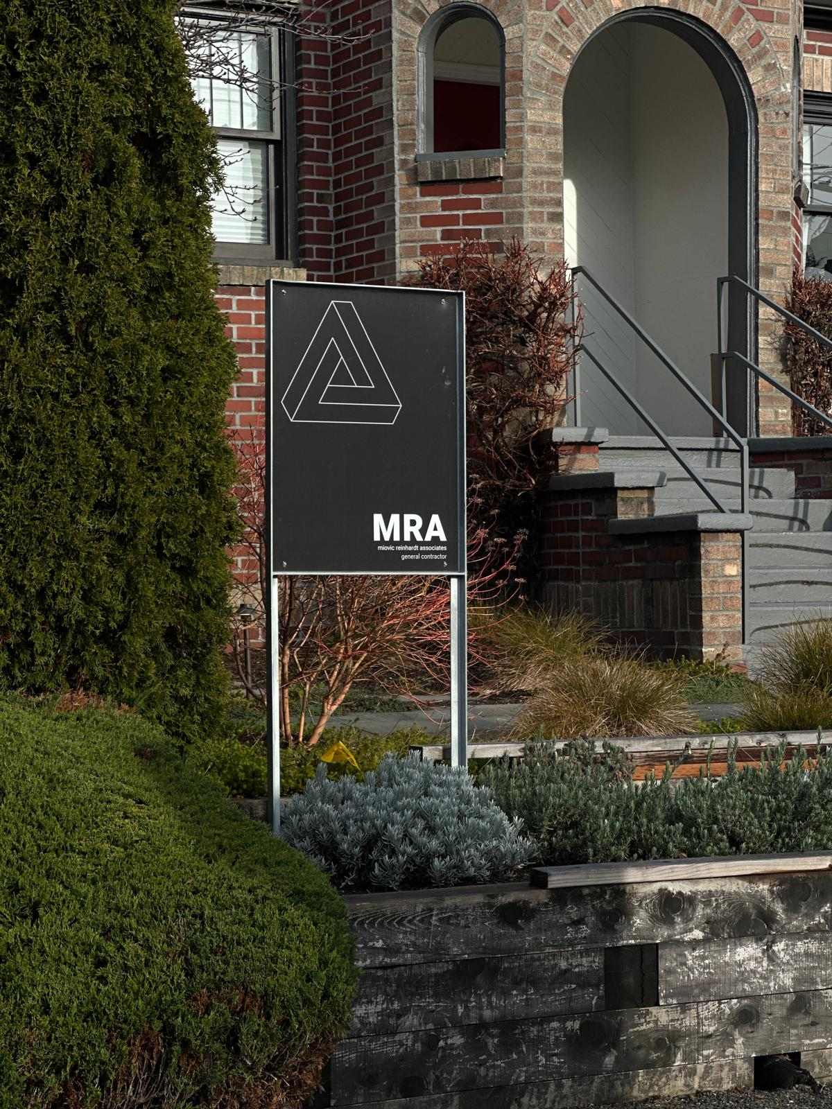 A black MRA sign with white triangle logo stands in front of a red brick house with arched entry.