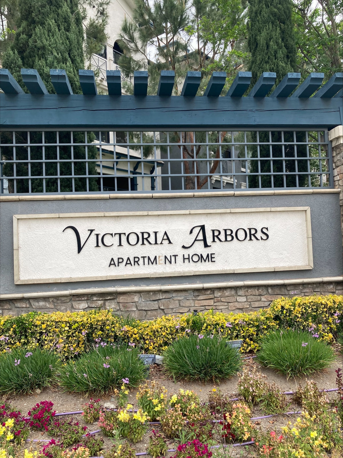 A Victoria Arbors apartment home sign mounted on a stone wall beneath a blue trellis with shrubs and flowers.