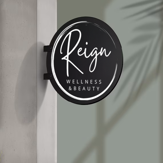 Black circular sign reading 'Reign' in script with 'Wellness & Beauty' below on a gray wall with palm shadow.