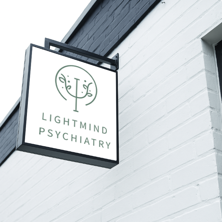 A hanging outdoor sign reads Lightmind Psychiatry with a green logo on a white panel against a white brick wall.