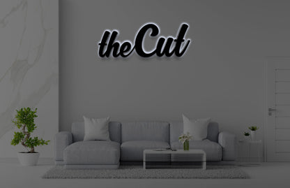 Modern gray living room with light gray sectional sofa, glass coffee table, potted bonsai, and black 'theCut' wall sign.