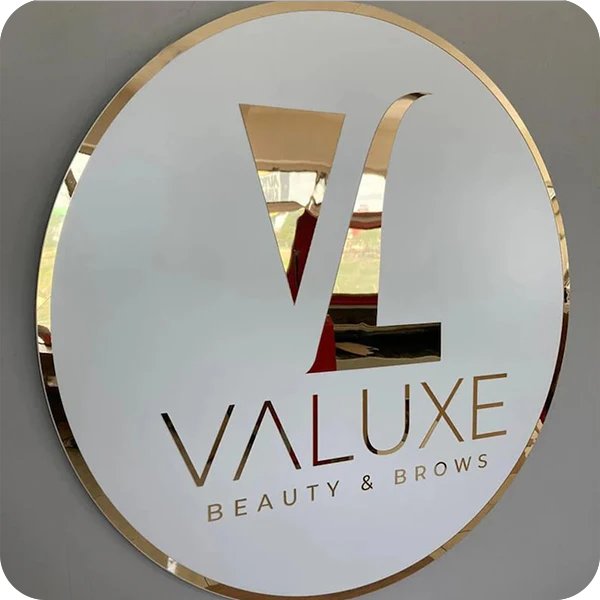 Round white sign with gold rim and reflective cutout 'VL' logo above gold 'VALUXE' and 'BEAUTY & BROWS' text.