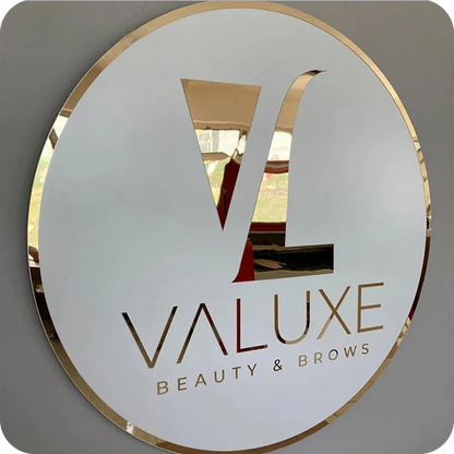 Round white sign with gold rim and reflective cutout 'VL' logo above gold 'VALUXE' and 'BEAUTY & BROWS' text.