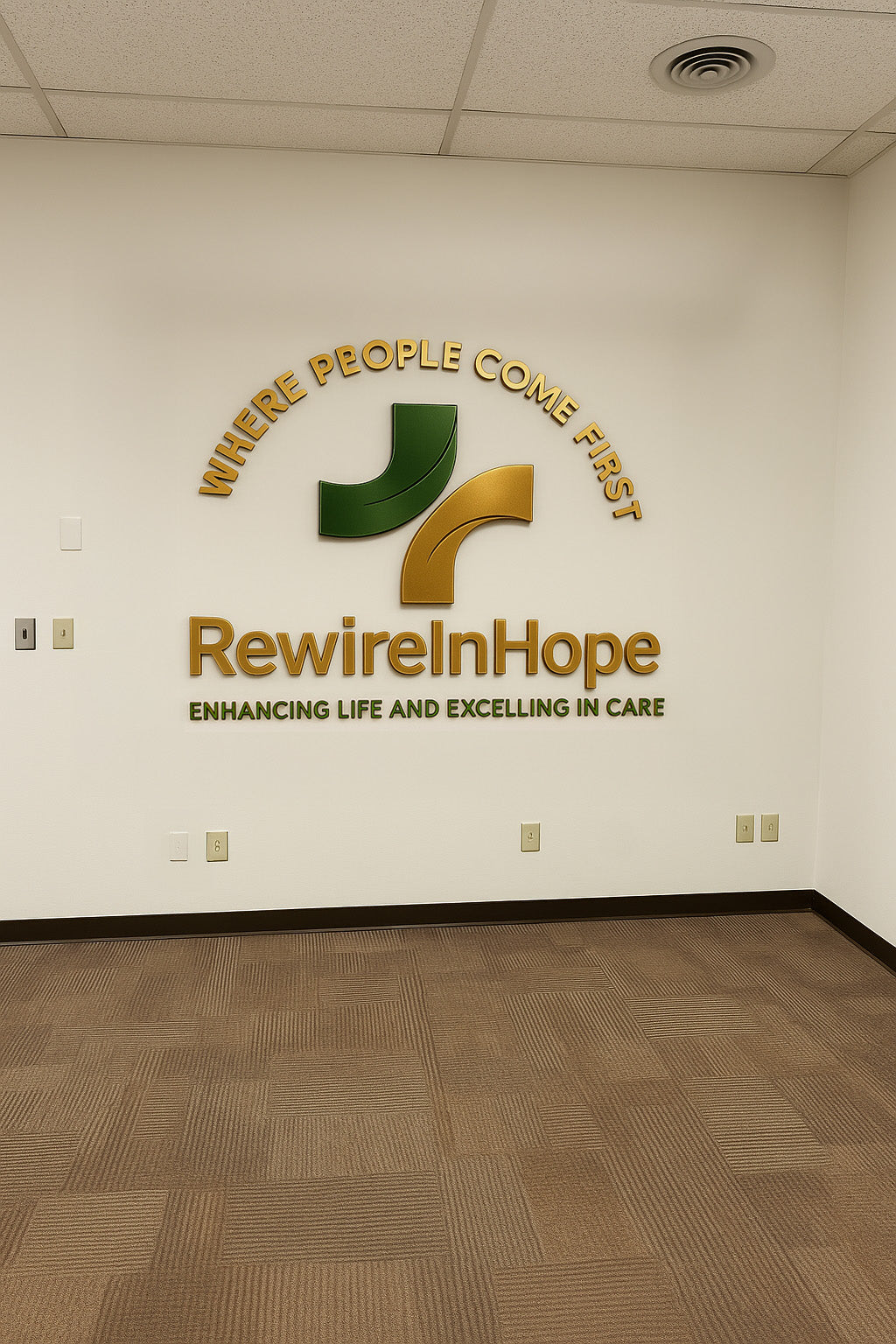 White office wall with a green and gold RewireInHope logo, 'Where People Come First' arch and tagline.